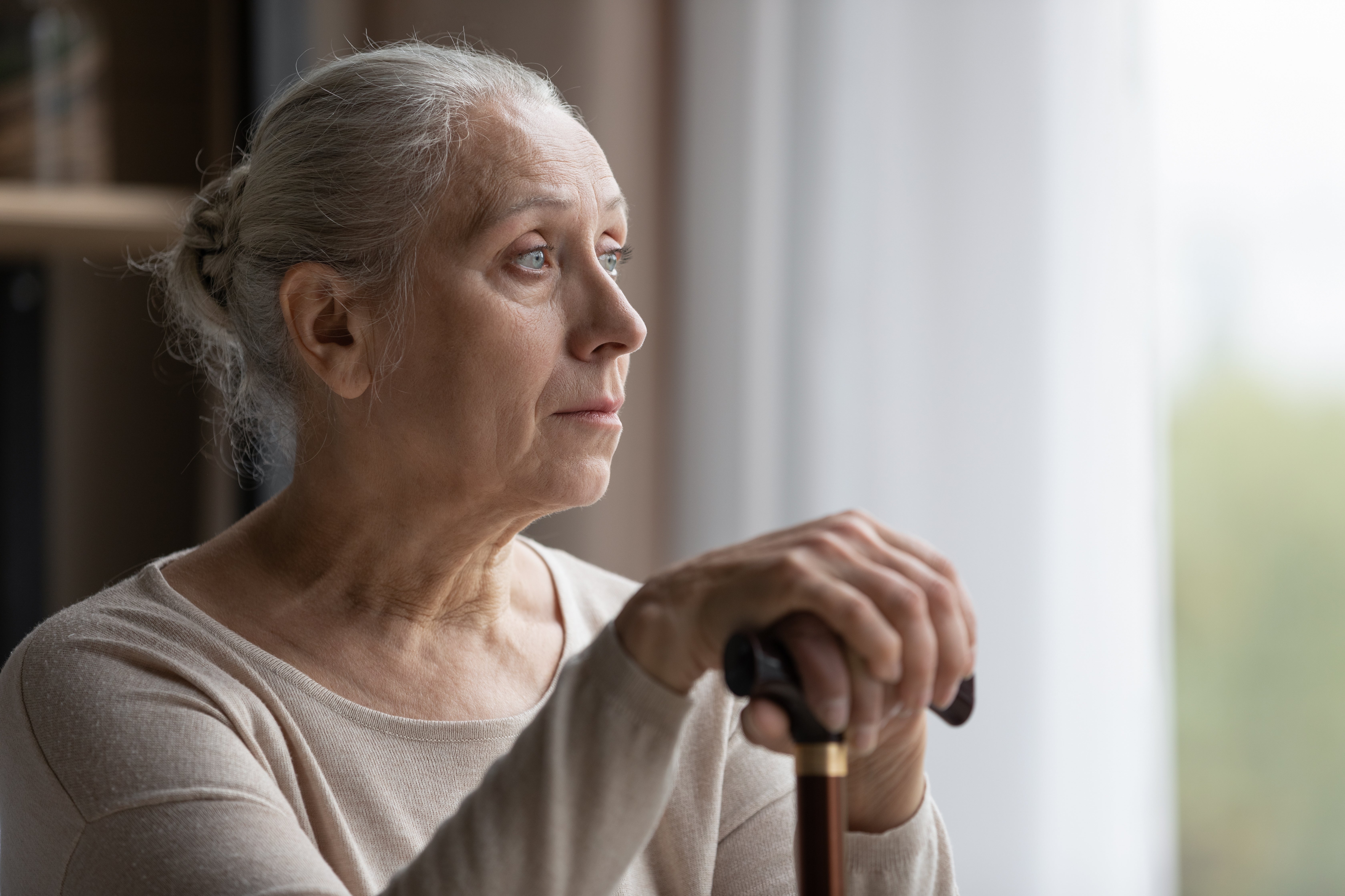 An older woman looks out the window with a downcast expression